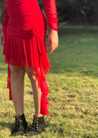 Woman wearing a red ruched dress with pleated hem and black boots, standing on grass