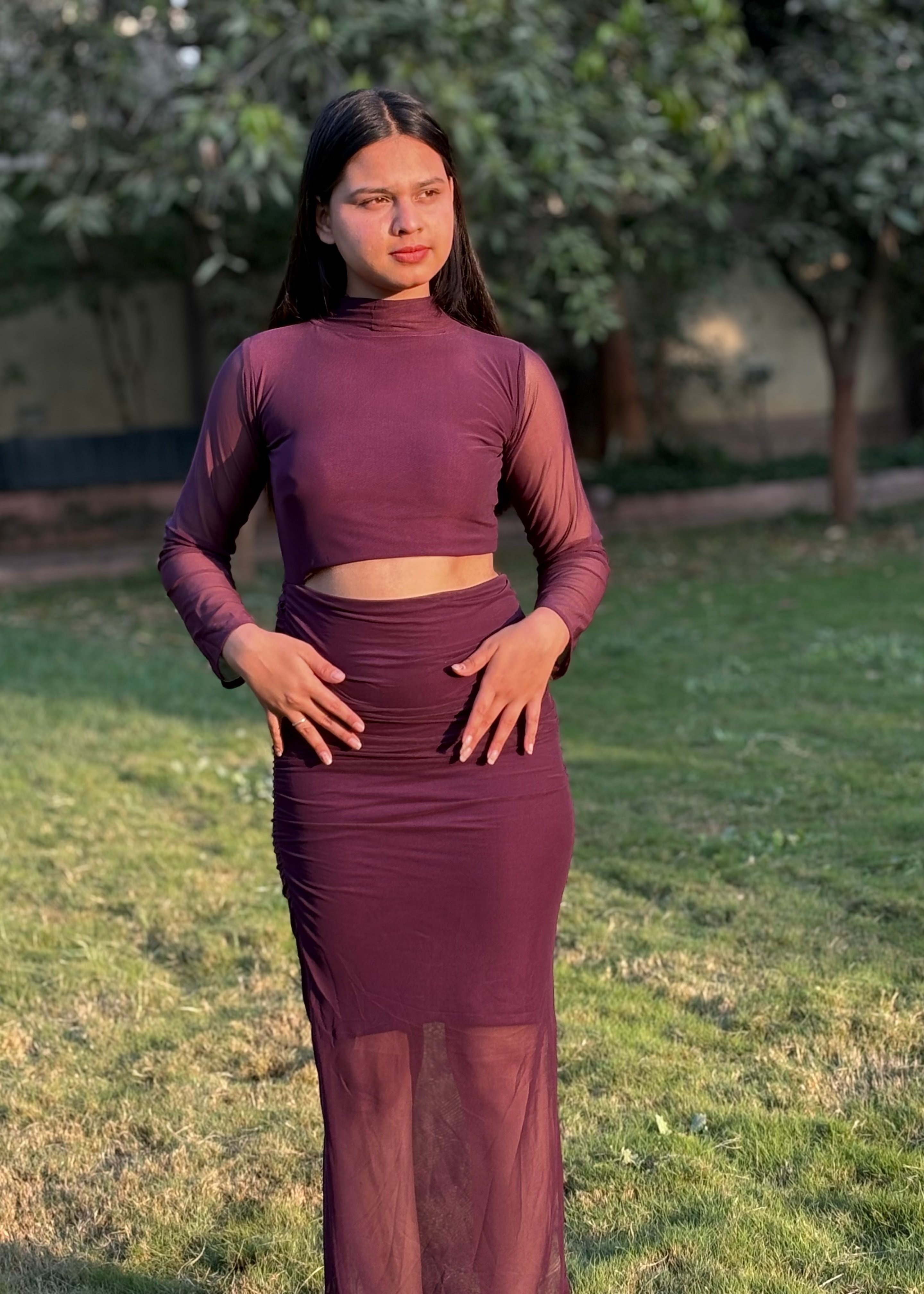 Woman in a fitted maroon sheer two-piece dress standing outdoors on grass