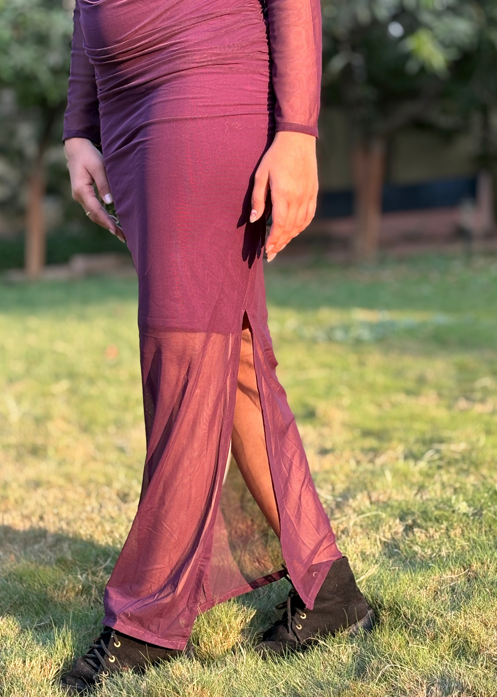 Woman wearing a sheer purple slit maxi dress and black boots, standing on grass