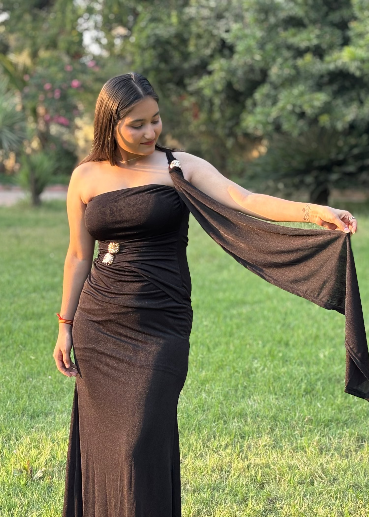 Woman in elegant black one-shoulder gown with draped sleeve, standing on green lawn