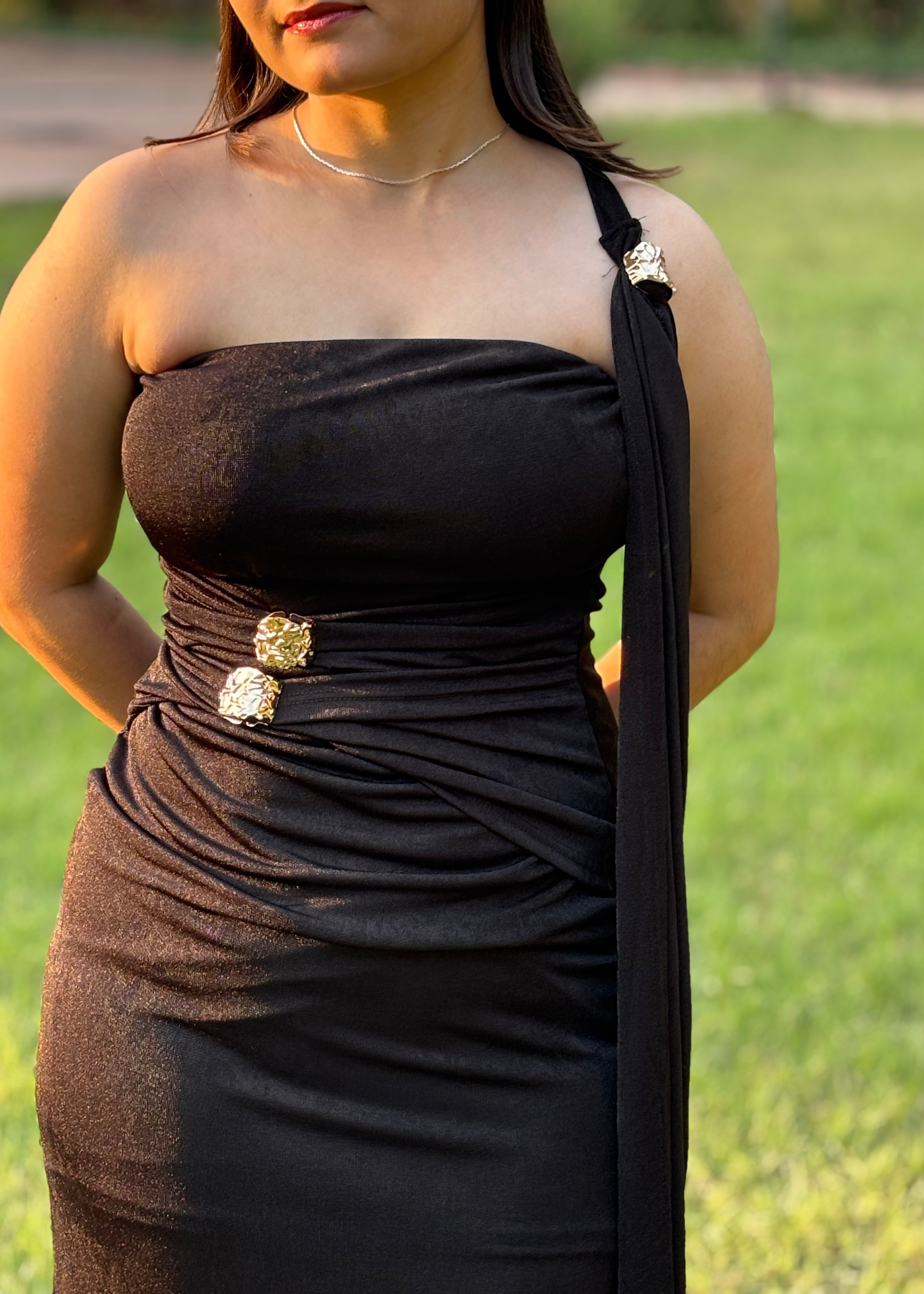 Woman in elegant black one-shoulder dress with gold embellishments, outdoors on grass