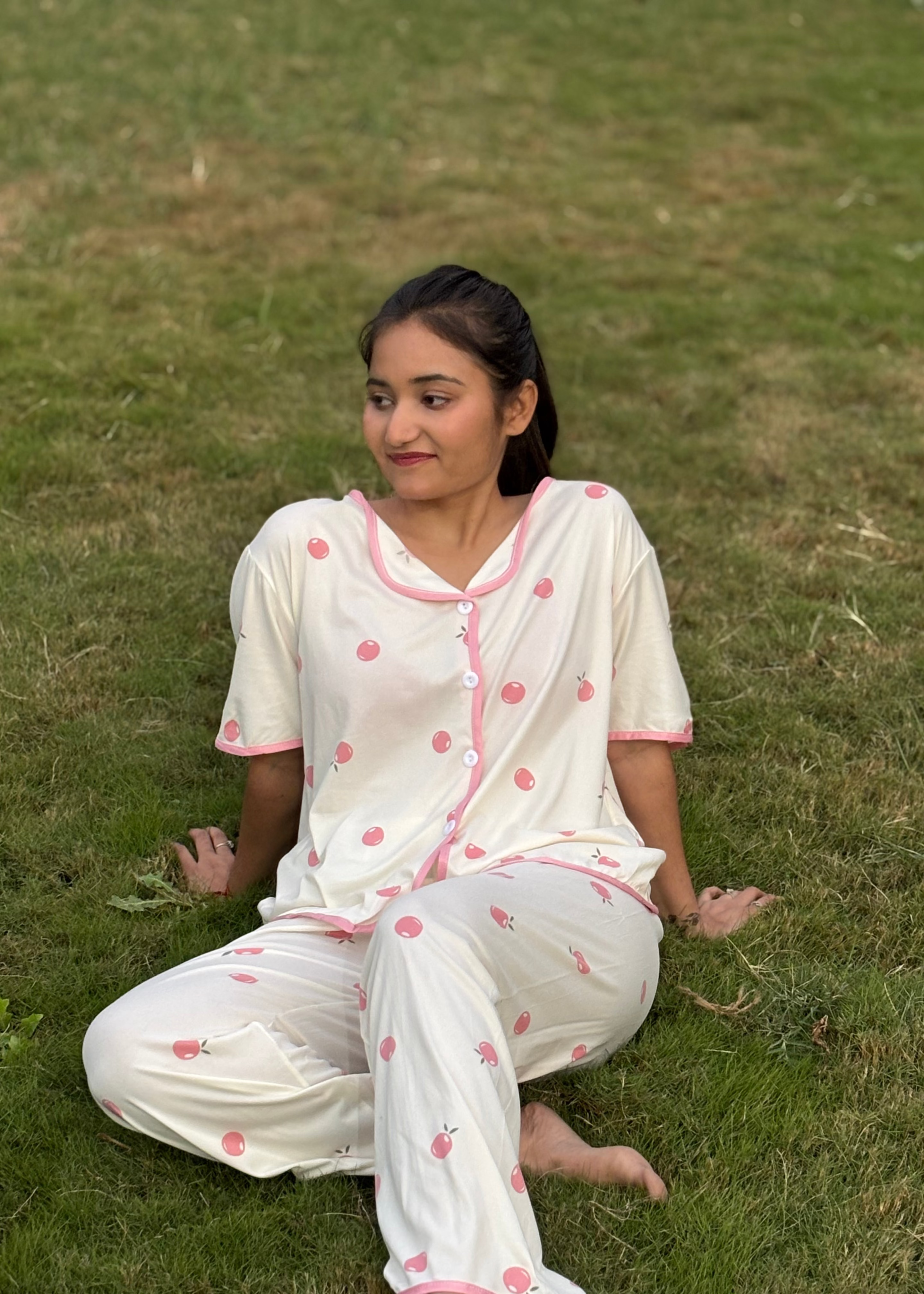 Woman sitting on grass in white cherry print pajama set with pink trim