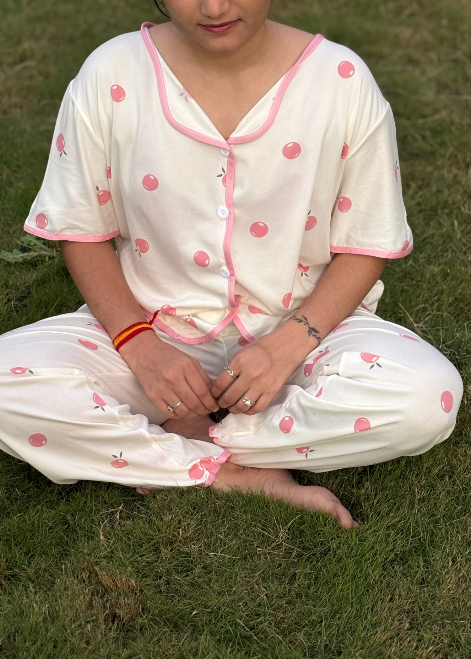 Woman in cream pajamas with pink cherry print sitting cross-legged on grass