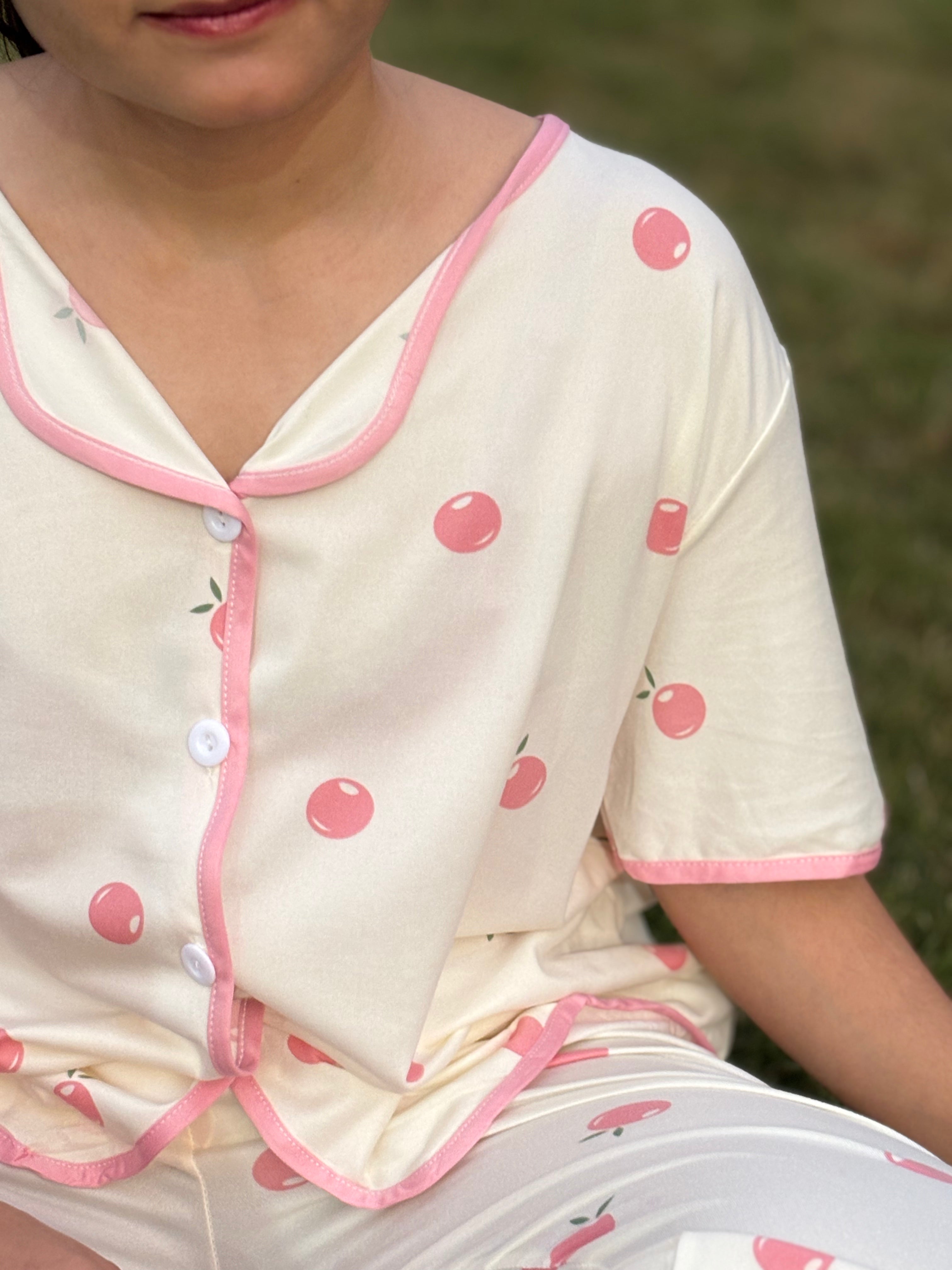 Girl wearing cream pajamas with pink cherry print and pink trim, sitting outdoors on grass