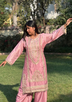 Woman wearing a pink embroidered kurta and trousers standing outdoors on green grass.